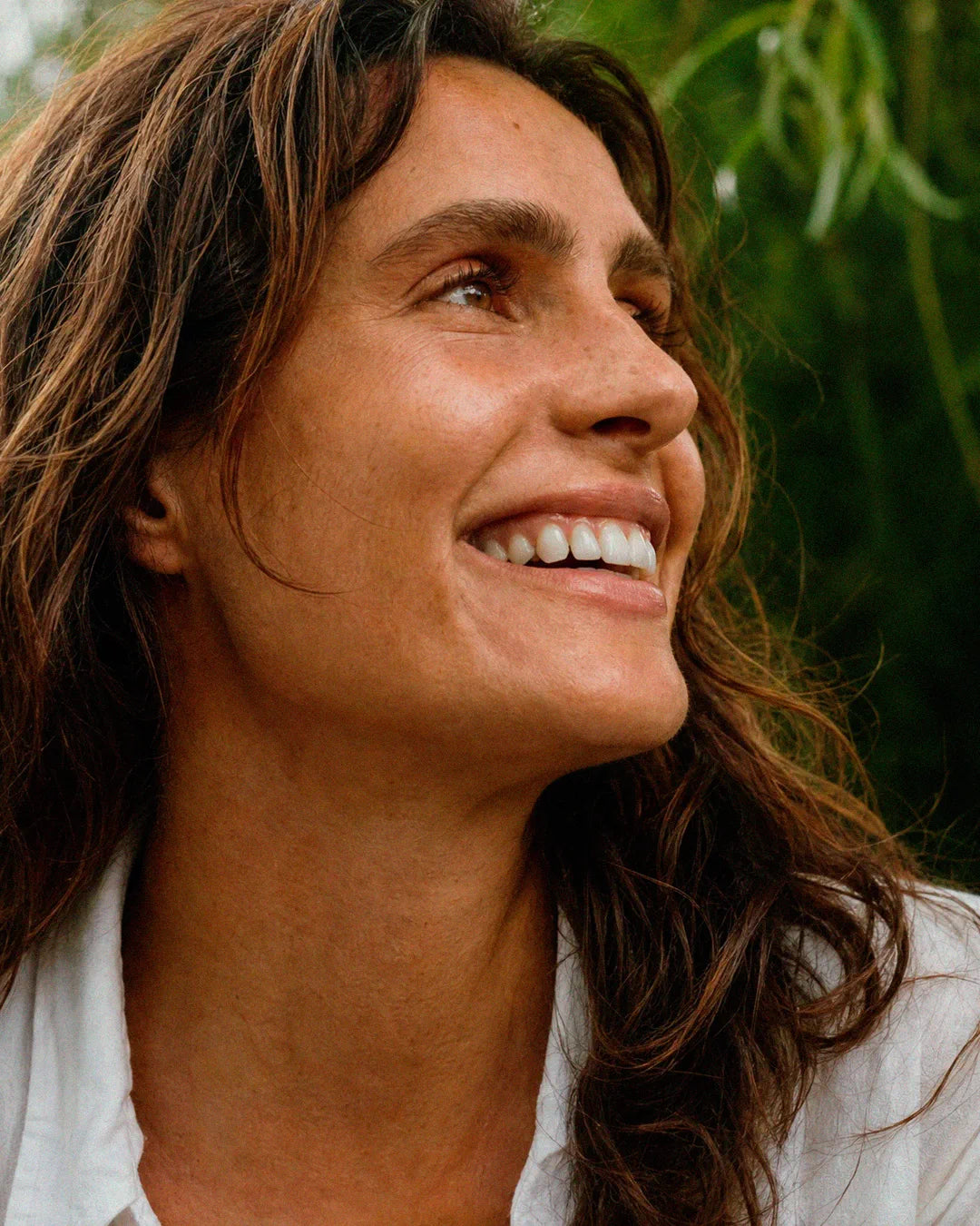 Mulher sorrindo ao ar livre, olhando para cima, com folhagem verde atrás
